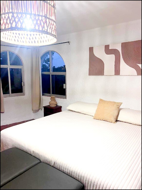 A well-lit sleeping area features a spacious bed with a textured white cover and a decorative pillow. Arched windows draw in natural light, framed by soft beige curtains. A minimalist wall art piece adds subtle color, while a bedside table holds a stylish lamp.