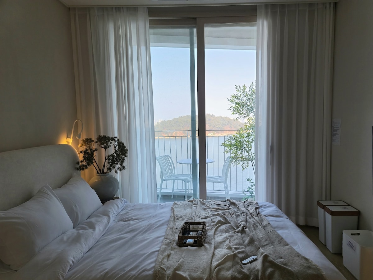 A serene bedroom space features a spacious bed topped with crisp white linens. Natural light fills the room through large glass doors, which open to a balcony with two chairs. Soft curtains frame the view of the sea and greenery outside.