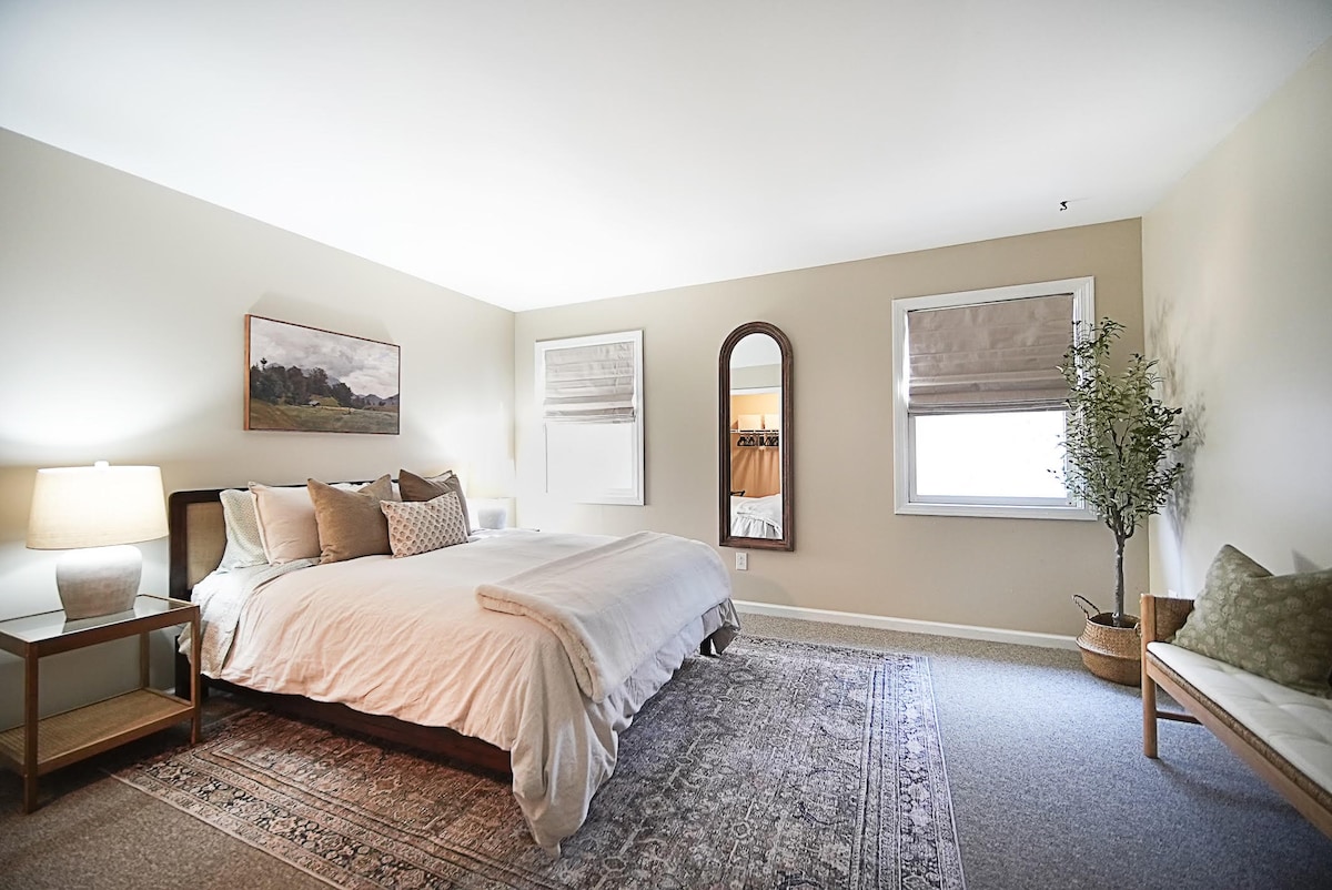 A spacious bedroom features a large bed dressed in soft linens, complemented by a bedside lamp on a wooden table. Natural light filters through a window covered by a light shade. A full-length mirror and an indoor plant add a touch of elegance to the decor.