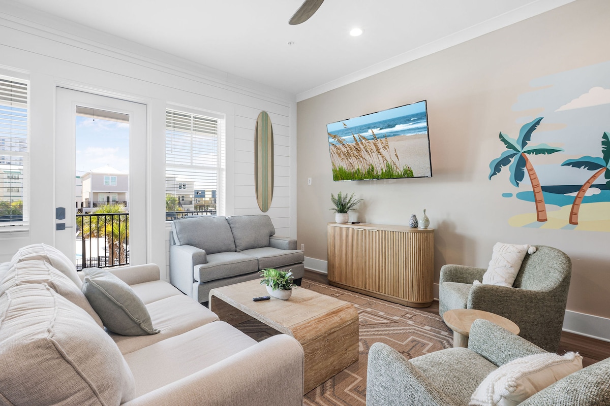 The living area features a spacious layout with a large sofa, love seat, and two comfortable chairs arranged around a wooden coffee table. A 65" smart TV is mounted on the wall opposite a decorative mural of palm trees against a soft backdrop.