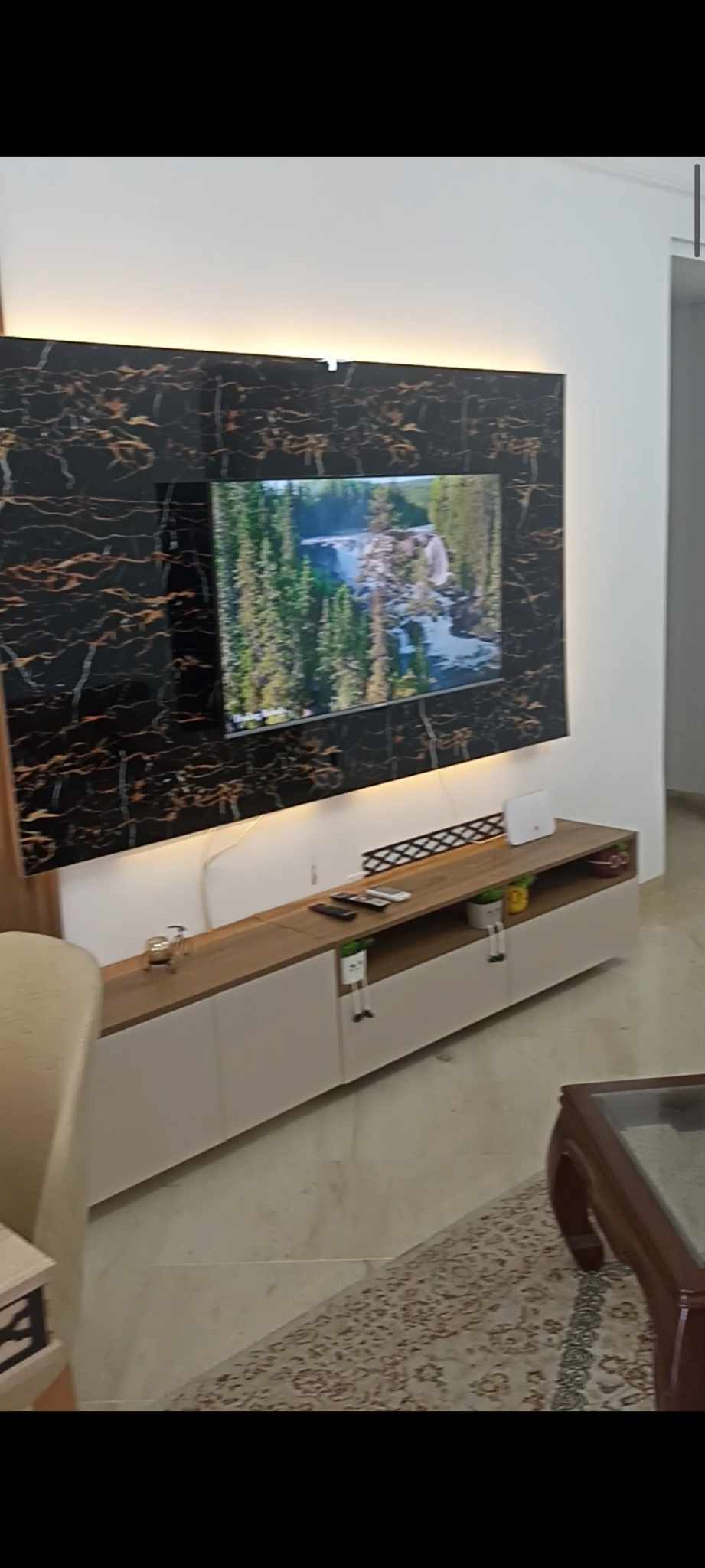 A modern living area is highlighted by a large wall-mounted television set against a stylish marble accent wall. Below, a sleek storage unit showcases a minimalistic design. Soft lighting enhances the space, while a cozy seating arrangement is visible in the foreground.