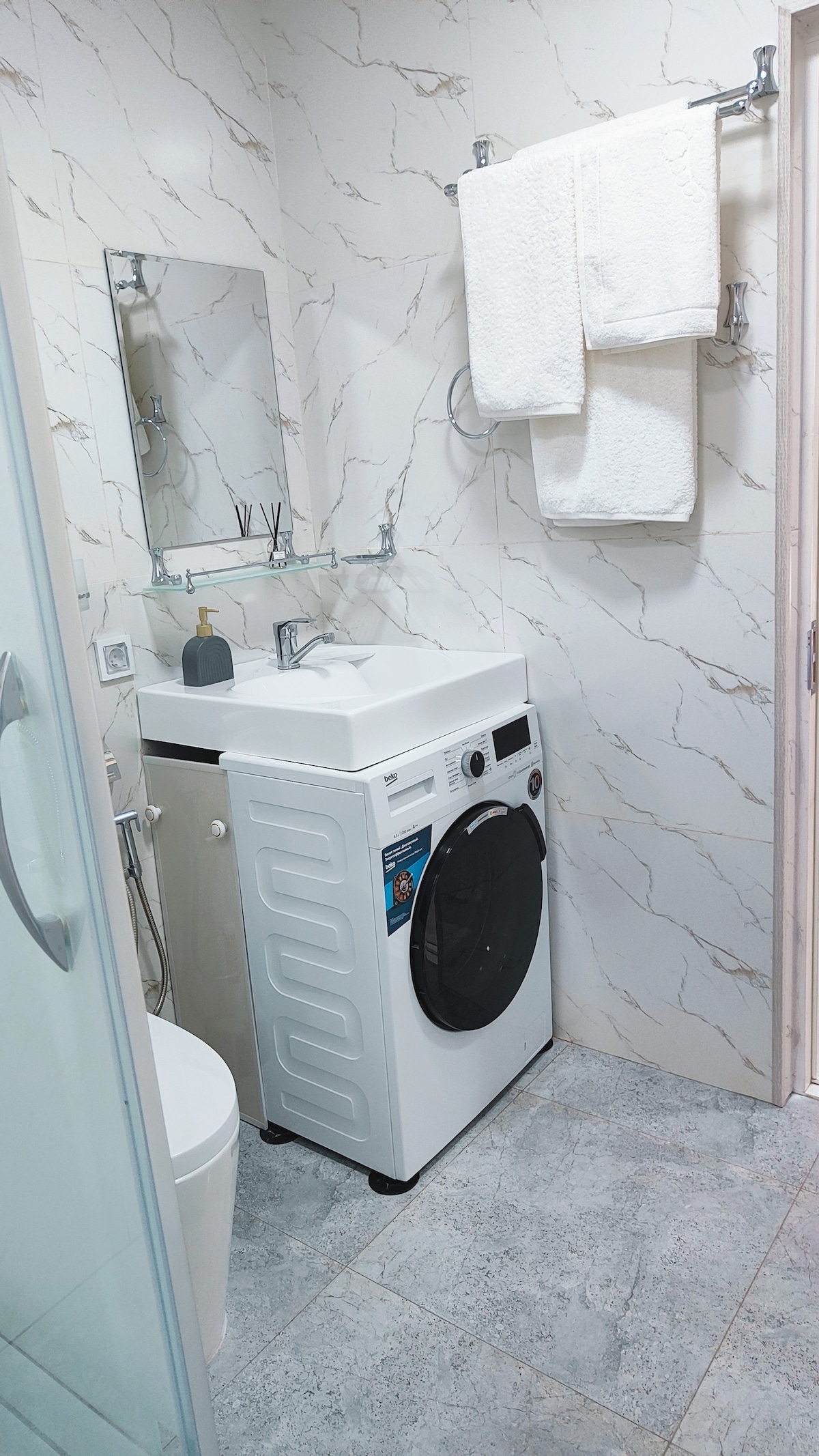 The bathroom features a sleek design with light-colored marble walls. A washing machine is positioned next to a modern sink, complemented by a mirror above. Fresh towels are hung neatly on a rack. The flooring is finished with gray tiles, contributing to the contemporary look.