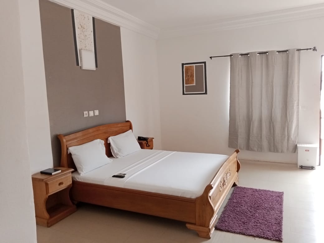 A well-furnished bedroom features a wooden bed with white linens, accompanied by a bedside table. A purple area rug lies on the floor, and a window dressed with light curtains allows natural light to brighten the room. Wall art adds a subtle touch to the decor.