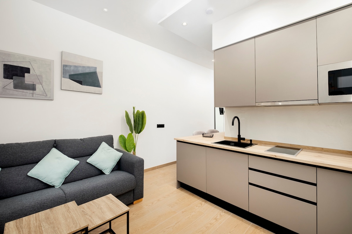 A modern kitchenette is paired with a cozy seating area featuring a gray sofa adorned with soft blue pillows. Two abstract artworks are displayed on the white wall, while a potted plant adds a touch of greenery to the space. Sleek cabinetry and appliances are visible.