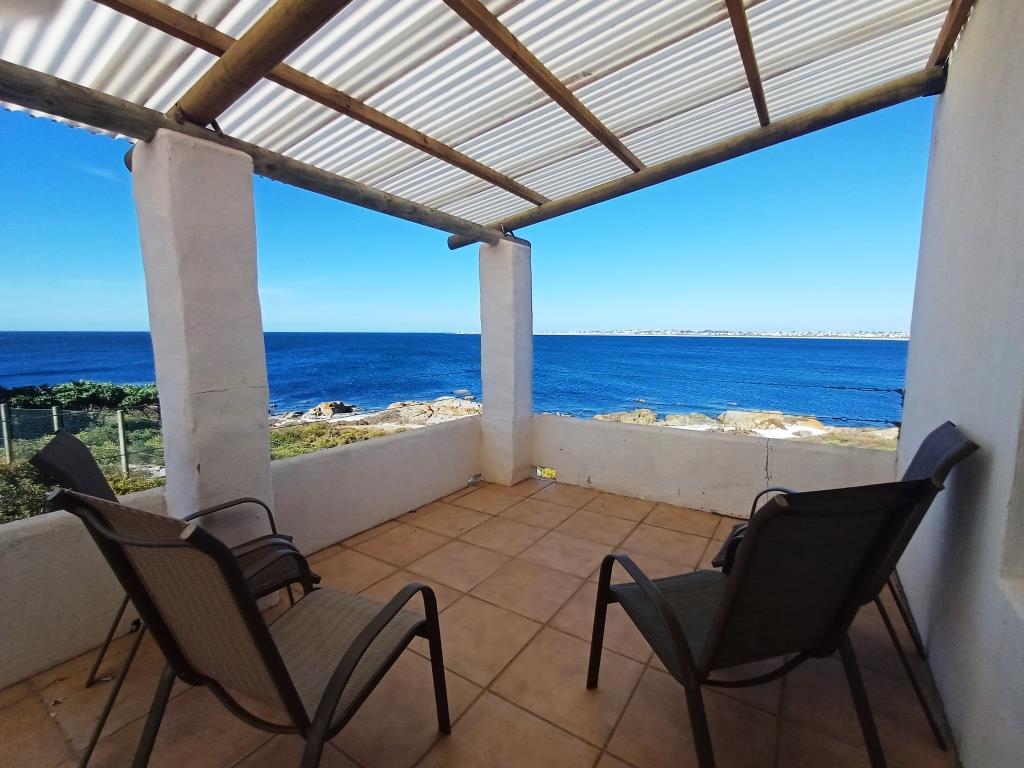 A shaded patio area features two lounge chairs positioned to overlook the ocean. The space is framed by sturdy white walls and a corrugated roof, enhancing the unobstructed sea view. The calming blue waters can be seen in the distance, providing a serene backdrop.