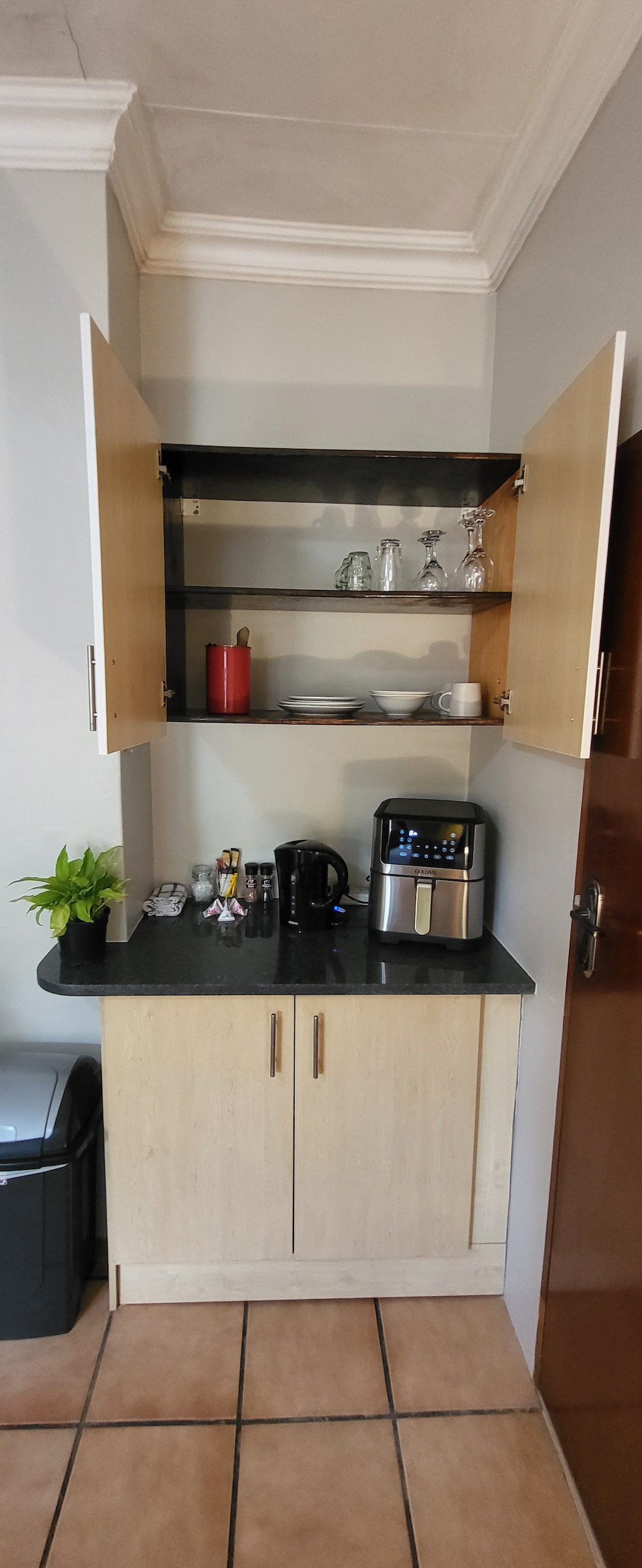 A kitchenette is depicted with open upper cabinets displaying glassware, dishes, and a decorative plant. A countertop holds a kettle and a coffee maker, complemented by a dark trash bin on one side. Ample storage space is provided below in the cabinetry.