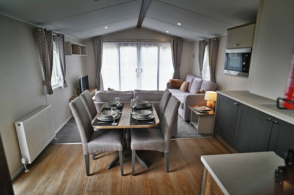 A modern lodge interior features a dining table set for four, complemented by stylish chairs. A cozy living area with a soft sofa is bathed in natural light from large windows, with curtains framing the view. A kitchenette is also visible in the background.