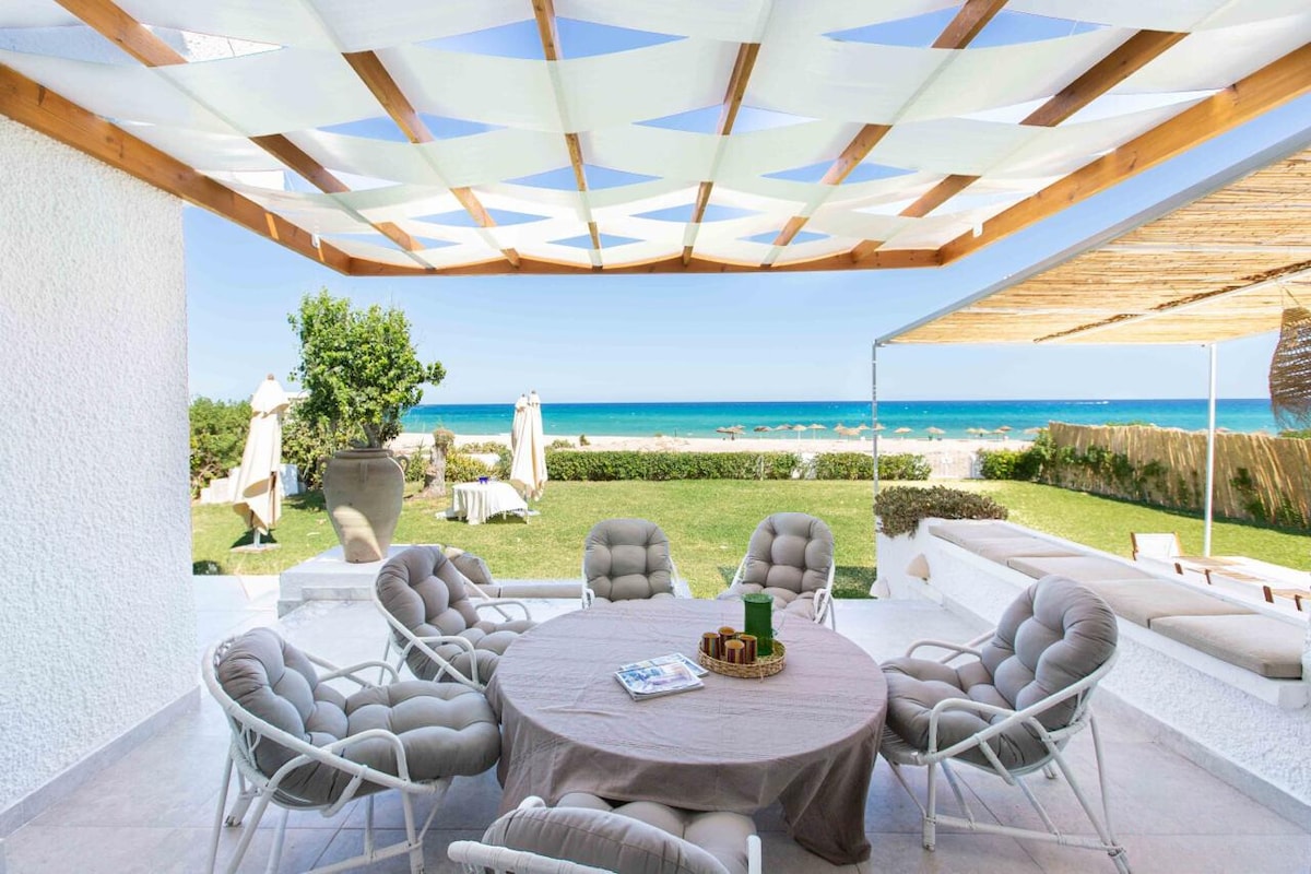 An outdoor dining area is featured under a shaded pergola, surrounded by six cushioned chairs arranged around a round table. Lush green grass leads to a view of the sea, while sun loungers can be seen in the background, emphasizing the serene coastal setting.