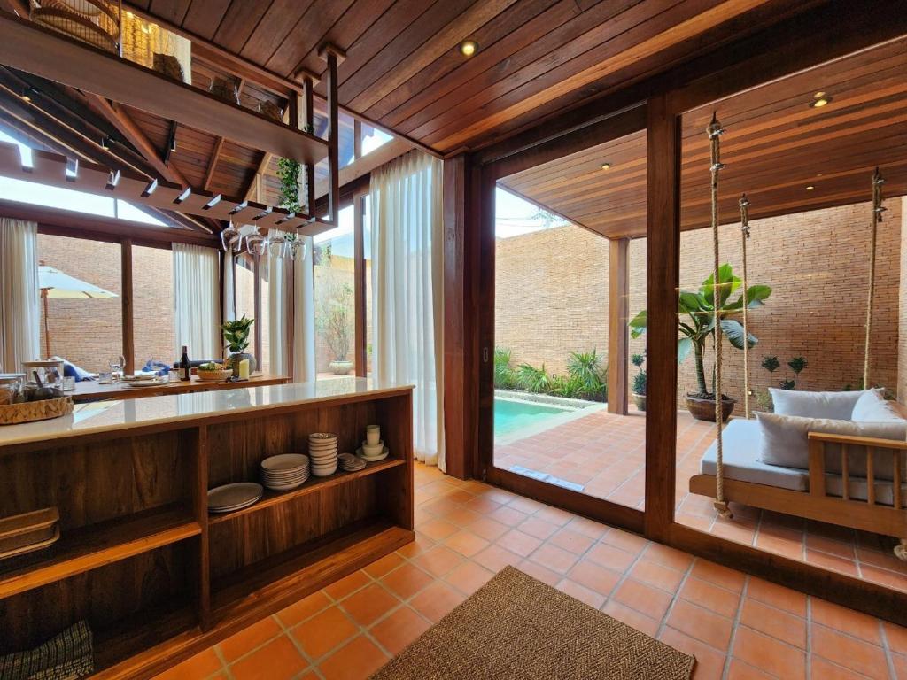 An open kitchen area is visible, featuring wooden cabinetry and a countertop. Floor-to-ceiling glass doors lead to a shaded outdoor space with a swimming pool and plants. A cozy seating area with cushions is seen, offering a relaxed atmosphere surrounded by natural light.