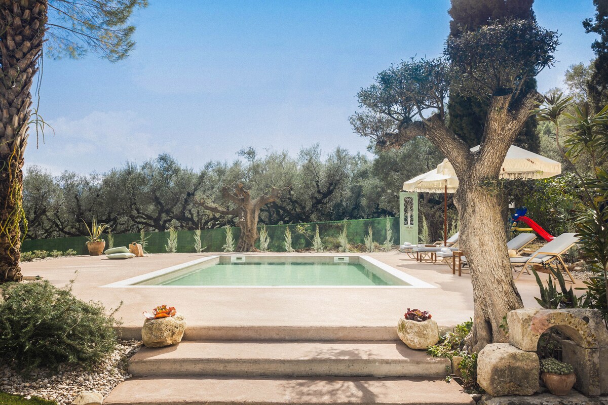 The image captures a serene pool area surrounded by lush greenery. A spacious pool is framed by chaise lounges and umbrellas, with olive trees dotting the landscape. Natural stone elements enhance the tranquil setting, inviting relaxation in a private outdoor oasis.