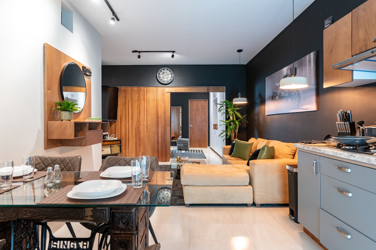 An airy living space features a glass dining table set for four, accompanied by a stylish sofa adorned with neutral-colored cushions. A large clock and wall art enhance the modern decor, while warm wood elements add a touch of sophistication. The kitchen area is visible, showcasing contemporary appliances.