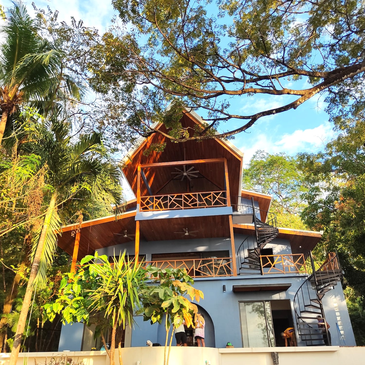 The exterior of a modern two-story house is shown, featuring a distinct angular roof and a spiral staircase leading to the upper level. Surrounding lush greenery and tropical trees enhance the tranquil setting. Large windows and a balcony are visible, inviting natural light into the space.