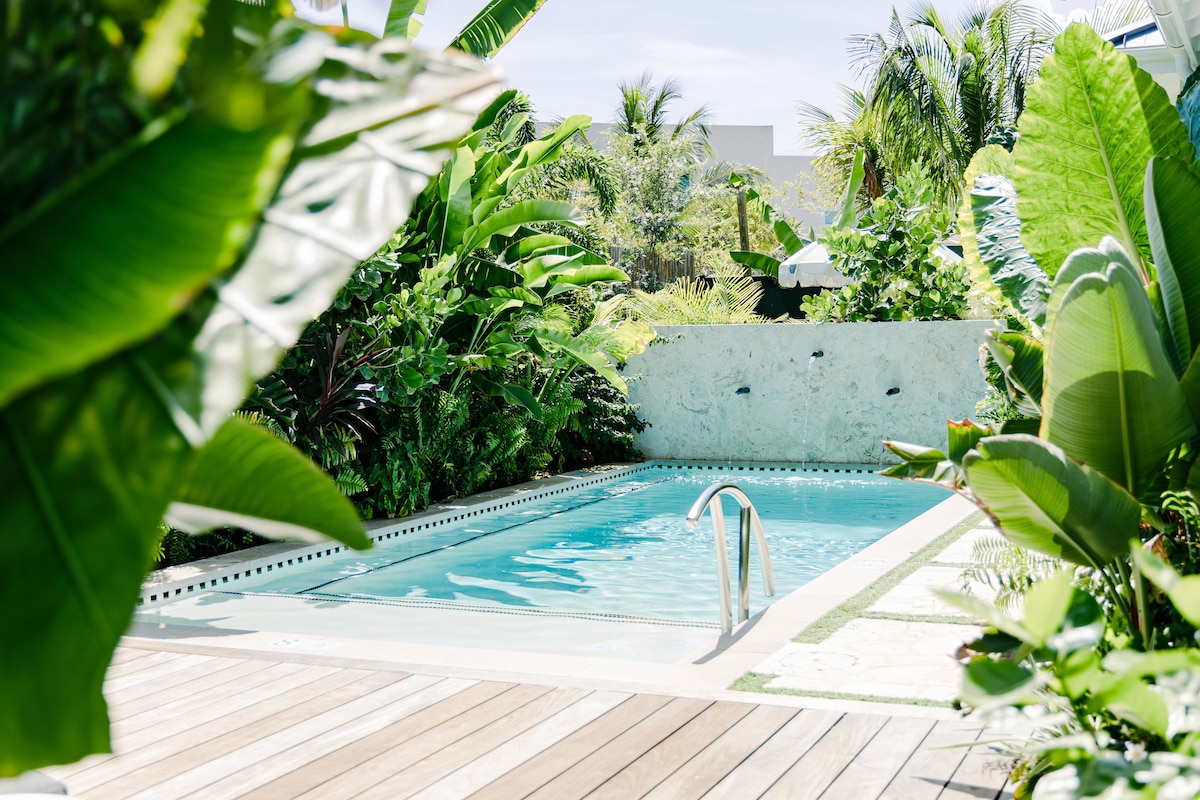 A serene swimming pool is framed by lush tropical foliage, with a gentle slope leading to the water. The pool's clear water reflects the surrounding greenery, while a wooden deck provides space for relaxation. Sunlight filters through the leaves, creating a calming atmosphere.