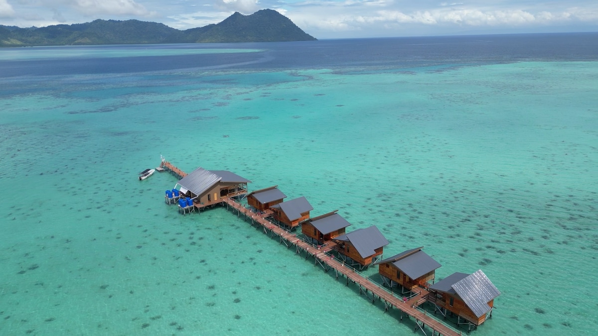An aerial view displays a series of wooden water chalets connected by a wooden walkway over vibrant turquoise waters. The surrounding sea extends towards distant green hills, emphasizing the remote tranquility of this unique seaside location.
