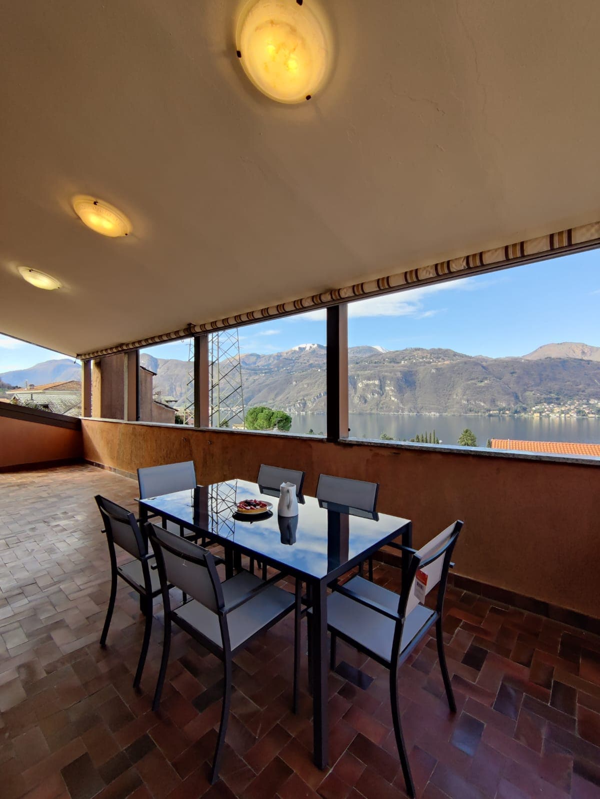 A covered terrace includes a dining table with six chairs, offering expansive views of the surrounding mountains and Lake Como. Large windows allow natural light to brighten the space, creating a pleasant environment for outdoor dining.
