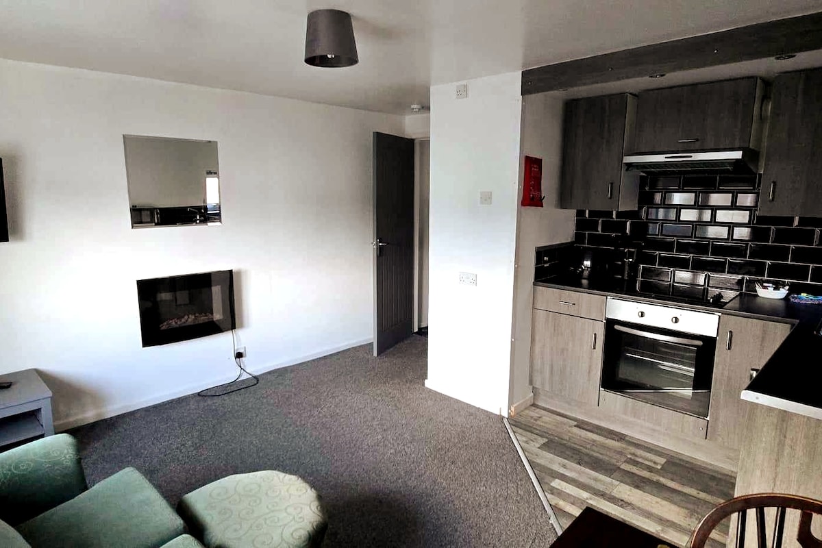 A living area is connected to a compact kitchen featuring modern appliances. A wall-mounted mirror reflects the space, while a cozy seating arrangement invites relaxation. Textured flooring enhances the overall layout, providing a comfortable setting for guests.