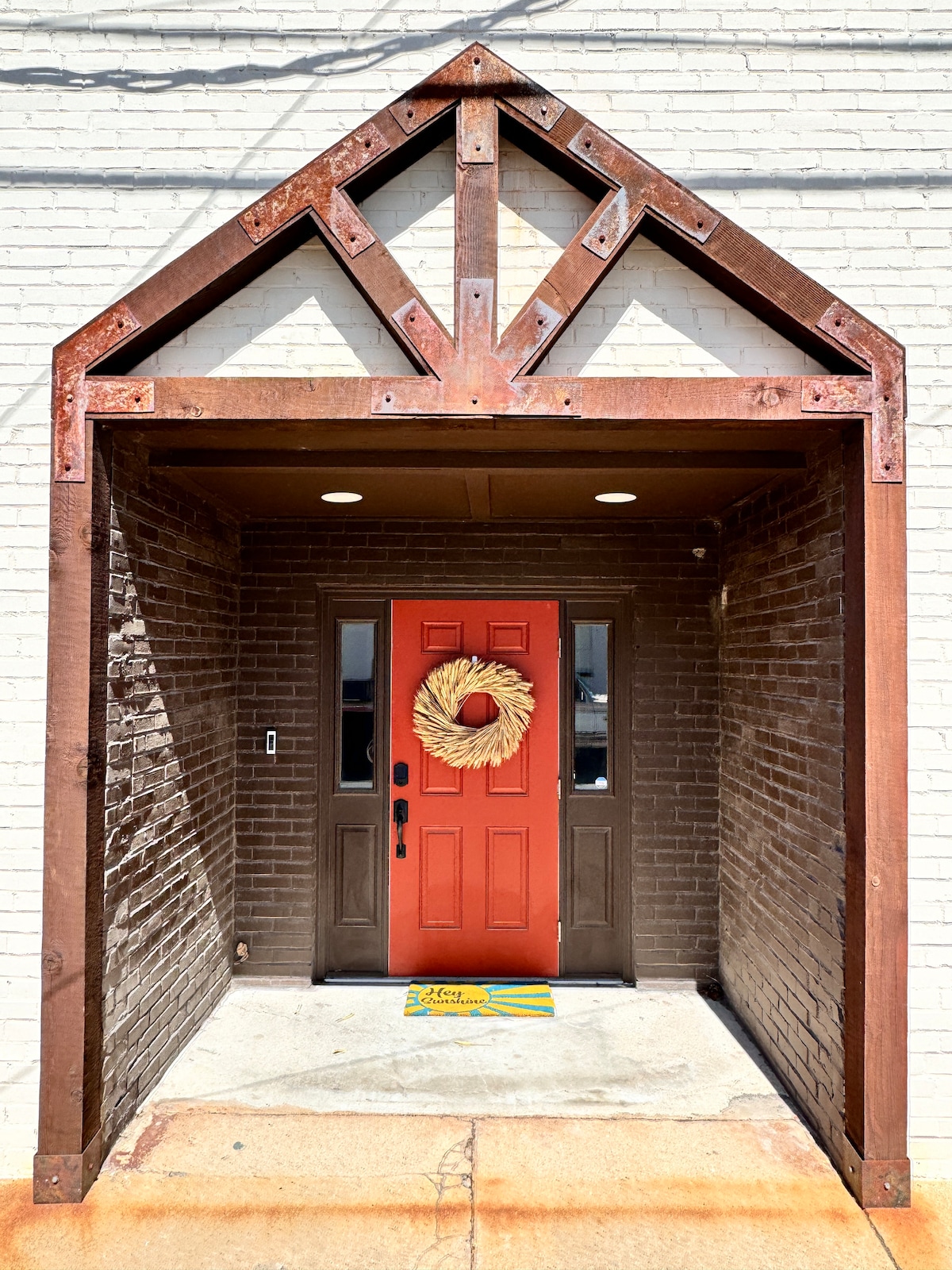 The entrance showcases a sturdy brick facade with a bold red door adorned with a wreath. A metal overhang highlights the architectural detail, while a welcome mat is positioned in front, inviting guests into the space.
