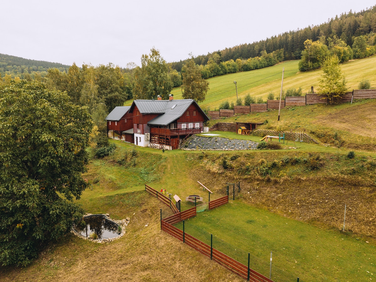 A rustic red wooden chalet is situated on a gently sloping hill, surrounded by green fields and trees. The exterior features a spacious deck, while the yard includes open space with a small pond and playground equipment in the background.