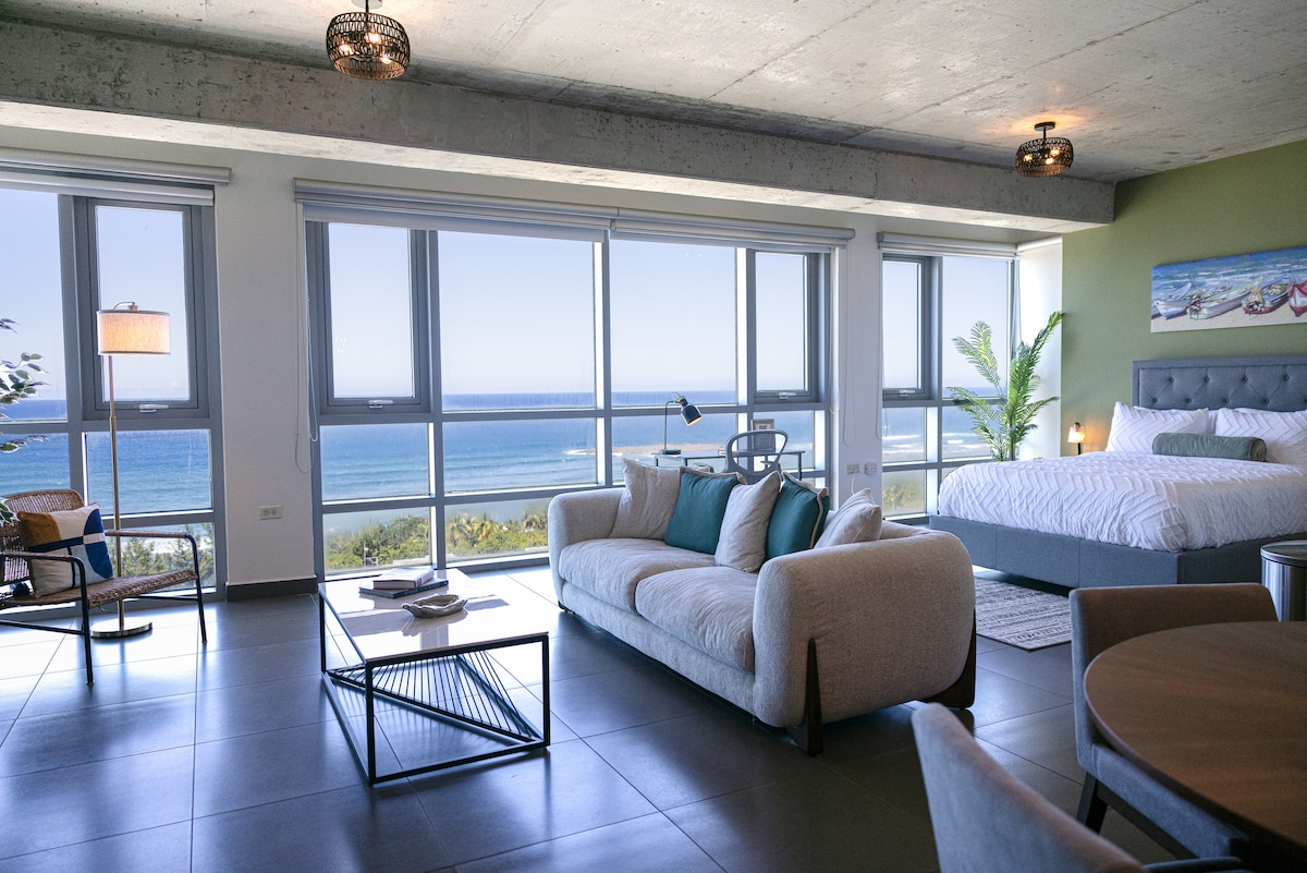 A spacious loft is shown with large windows offering a beautiful view of the Atlantic Ocean. A cozy seating area is visible with a sofa and coffee table, alongside a round dining table. A queen size bed is neatly made in the corner, complemented by modern decor.