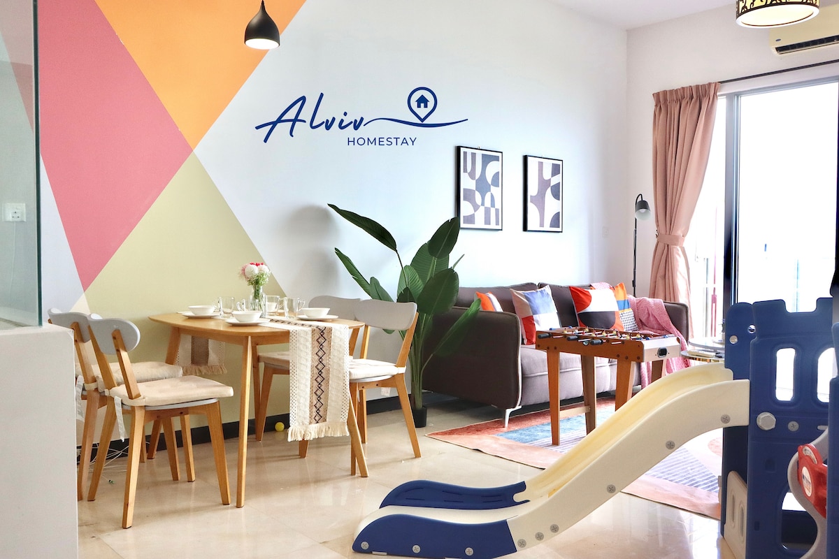 The living area features a dining table with light wooden chairs, complemented by a colorful wall design. A comfortable sofa and a small games area are visible. A potted plant adds freshness, while large windows provide natural light. The space is family-friendly.