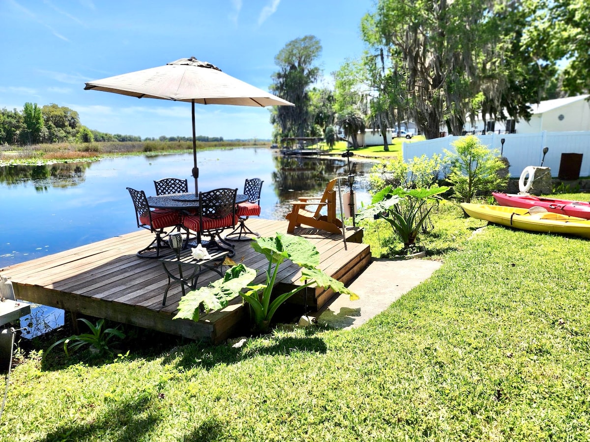 A lakeside deck features a large umbrella providing shade over a dining table with several chairs. Nearby, two kayaks are positioned on the grass, and lush greenery surrounds the area, creating a peaceful outdoor setting. A calm water view reflects the surrounding nature.