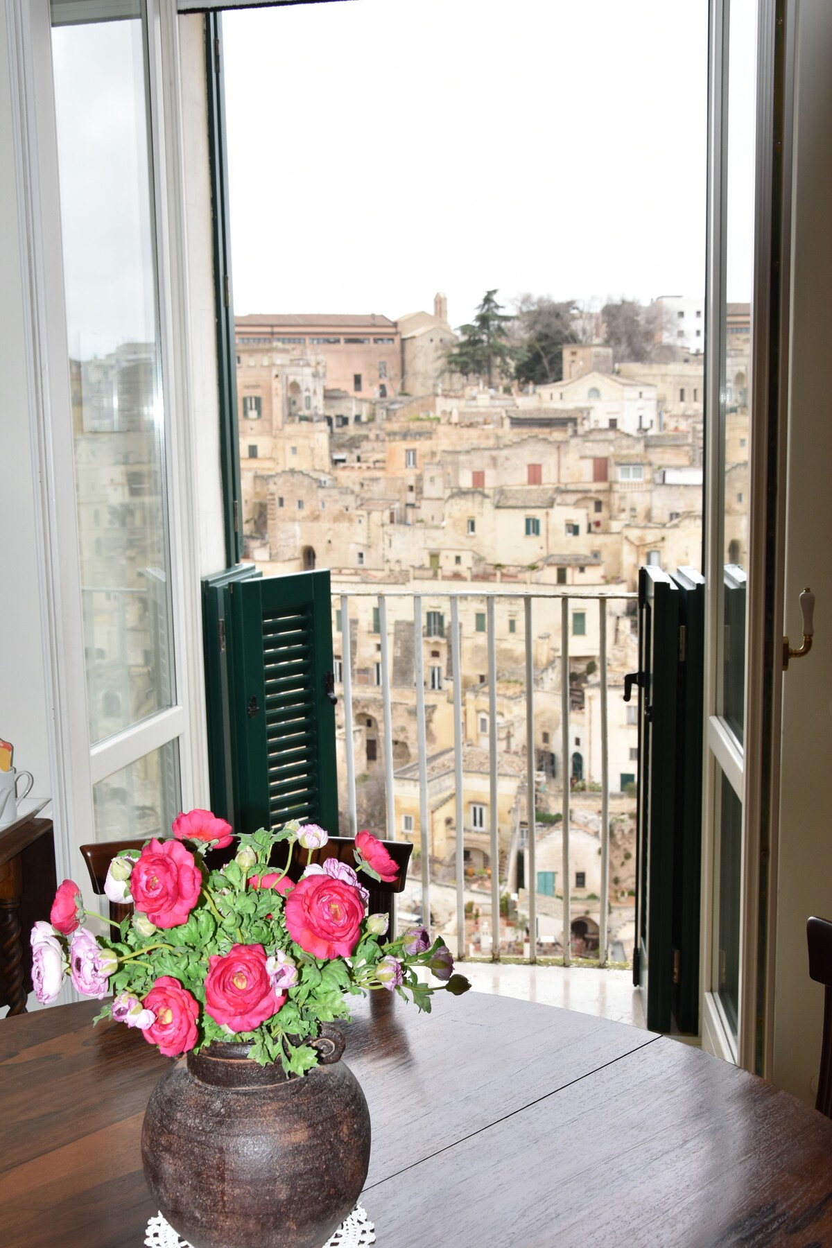 A view through open doors reveals picturesque stone buildings of the historic Sassi district. A wooden table is adorned with a vase of pink flowers, complementing the natural light filtering in from the balcony.