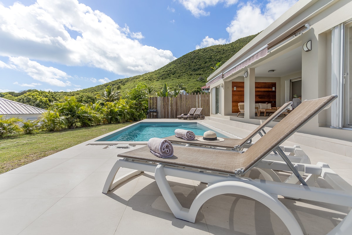 Sun loungers are positioned beside a clear pool, offering views of a lush, green hillside. The exterior design shows large sliding glass doors that open to a spacious living area, complemented by well-maintained landscaping.