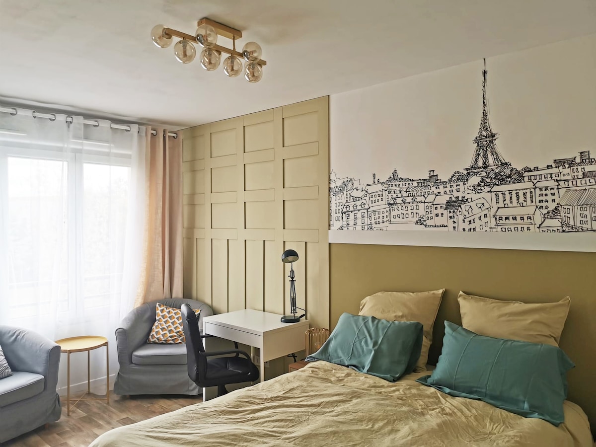 A bright and spacious room features a large bed with soft pillows and a light duvet. A minimalist desk and chair are positioned against a wall adorned with a line drawing of the Eiffel Tower. Two comfortable armchairs and a small table complement the layout.