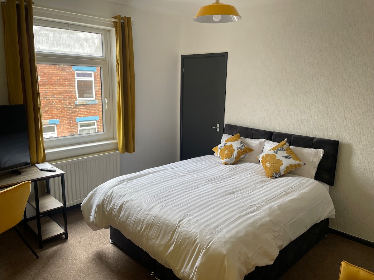 A spacious bedroom is furnished with a large bed adorned with white linens and decorative yellow pillows. A television stands against one wall, while a chair is placed beside it. Natural light enters through a window, framed by yellow curtains.