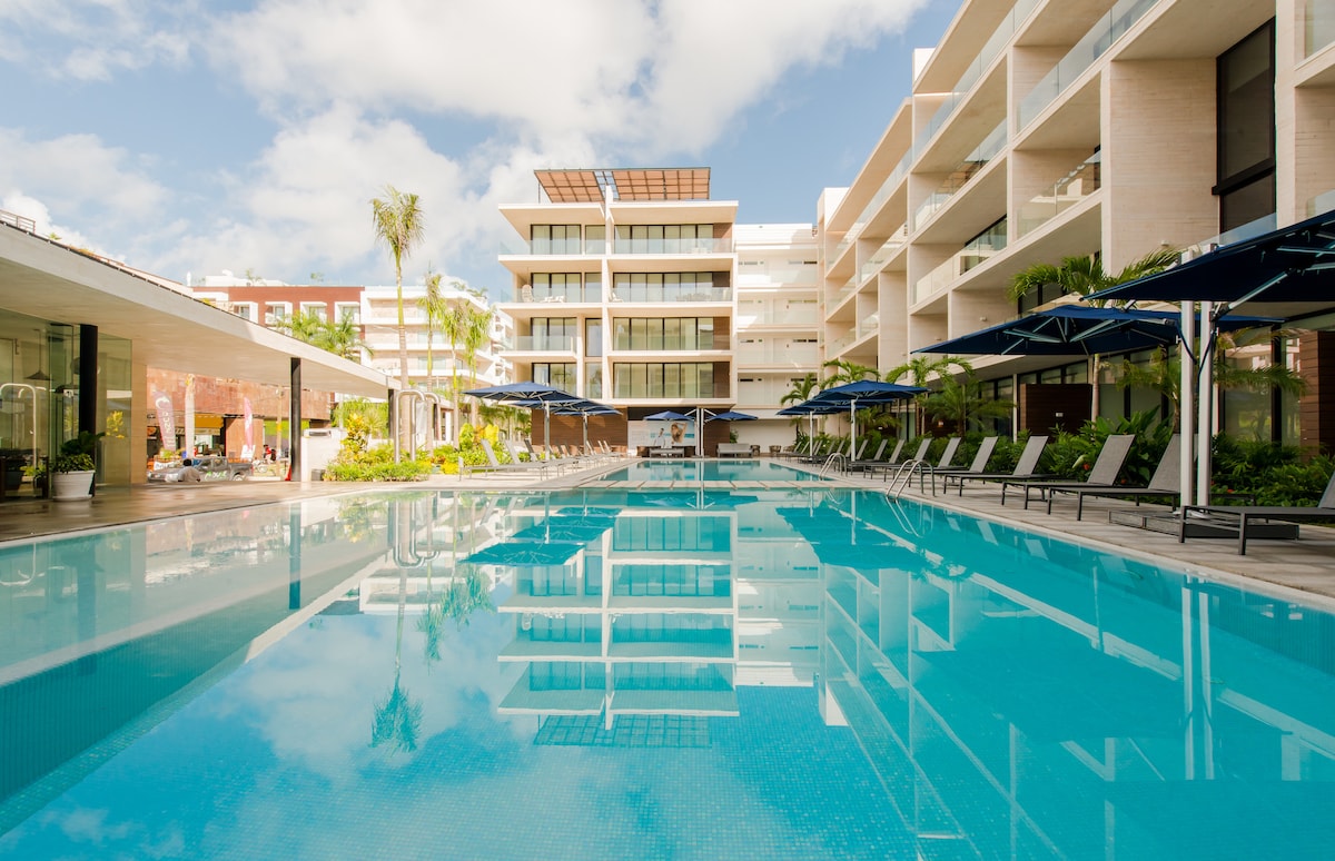 A serene pool area is showcased, featuring clear blue water that reflects the surrounding buildings and lush greenery. Lounge chairs under large umbrellas line the poolside, inviting relaxation in a tranquil outdoor space.
