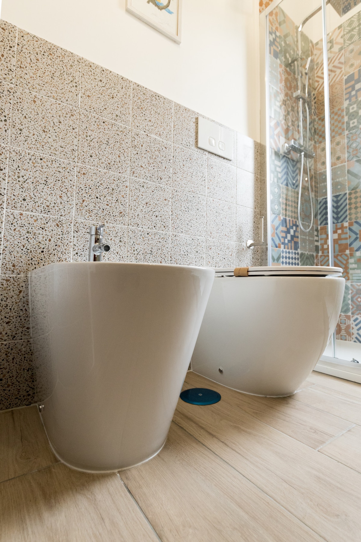 A modern bathroom features a stylish bidet and toilet with sleek, curved designs. The wall is adorned with textured tiles in geometric patterns, while the shower area is enclosed with clear glass, allowing natural light to illuminate the space.
