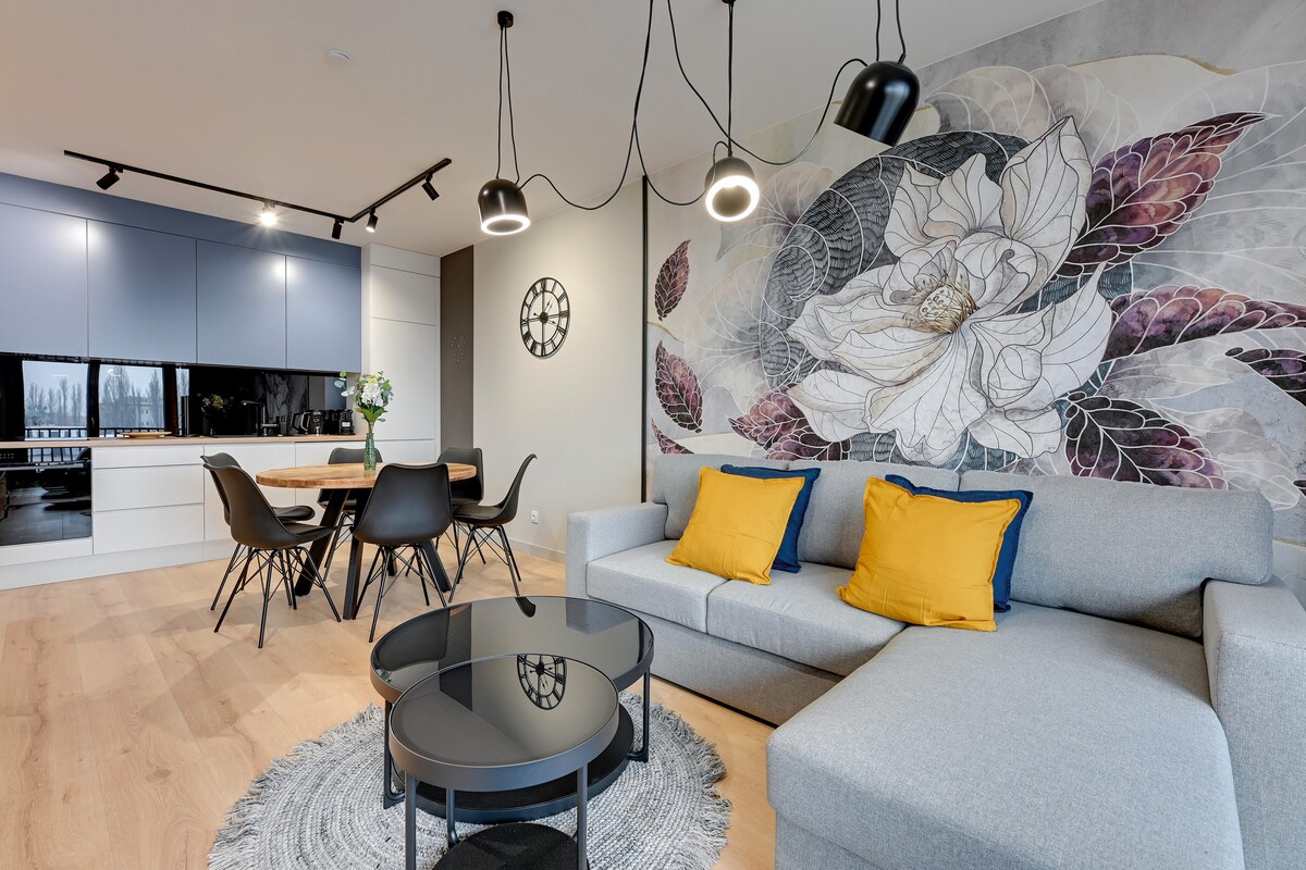 A modern living space features a light gray sofa adorned with yellow cushions. A round black coffee table is centered on a decorative rug. The wall behind the seating area displays a large floral mural. A dining table, accompanied by black chairs, is positioned in the background.