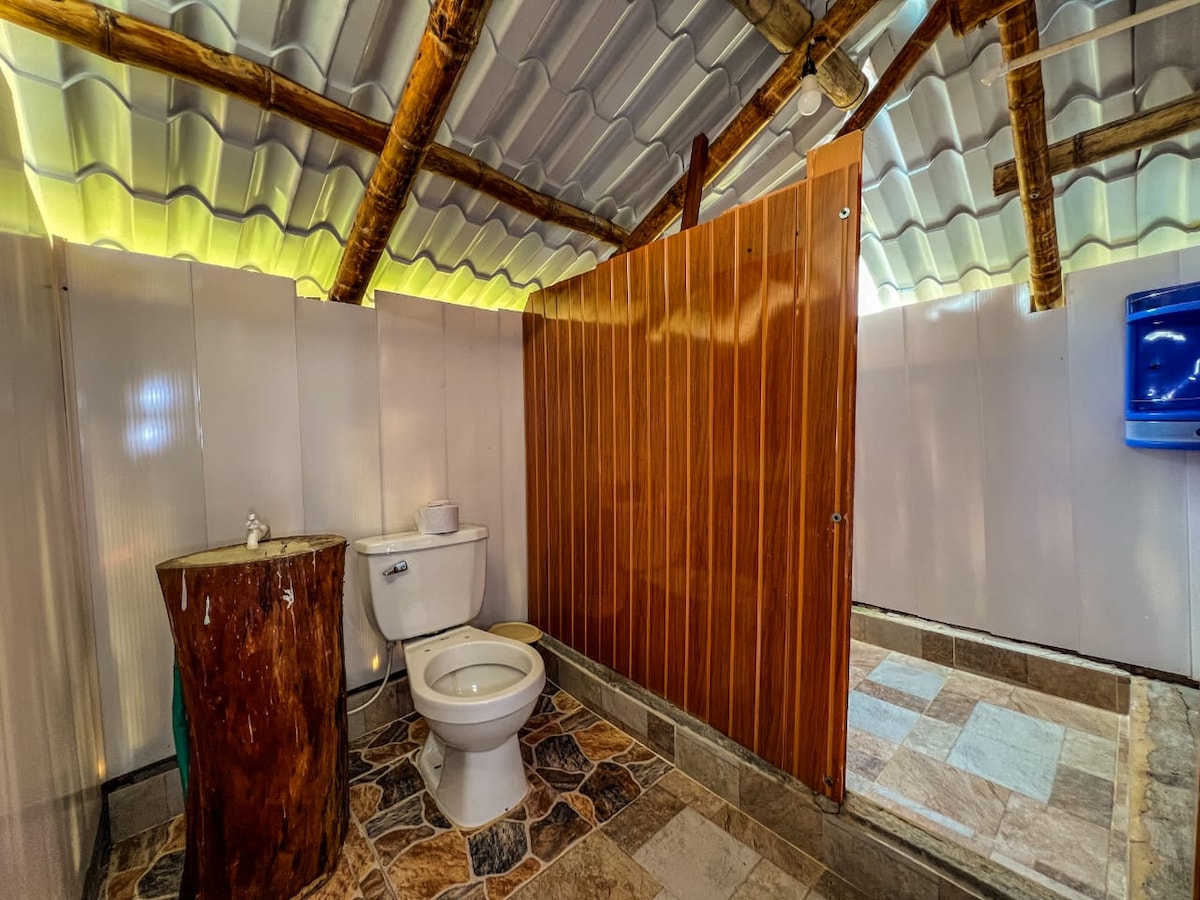 A spacious bathroom is showcased, featuring a wooden partition and a modern toilet. The flooring is composed of stone tiles, and natural light filters through the ceiling, highlighting the clean and simple design. A small wooden table is positioned nearby.