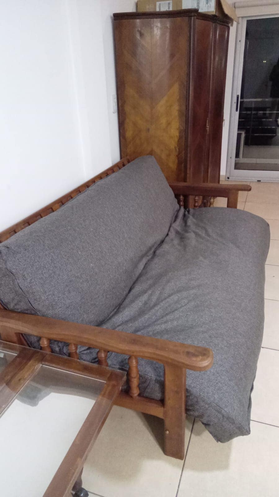 A wooden-framed sofa is featured, upholstered in a soft gray fabric. It is accompanied by a glass coffee table positioned nearby. Large sliding doors can be seen in the background, leading to an outdoor area.