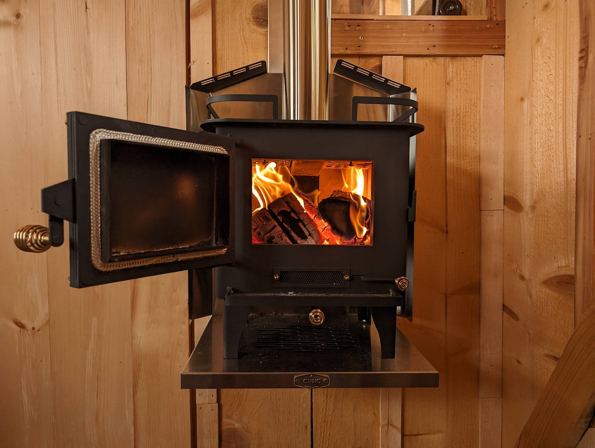 A small cubic wood stove is positioned within a wooden interior. The door is open, revealing a lively fire inside, casting warm light on the surrounding wood paneling. A surface beneath the stove provides a practical space for placing items.