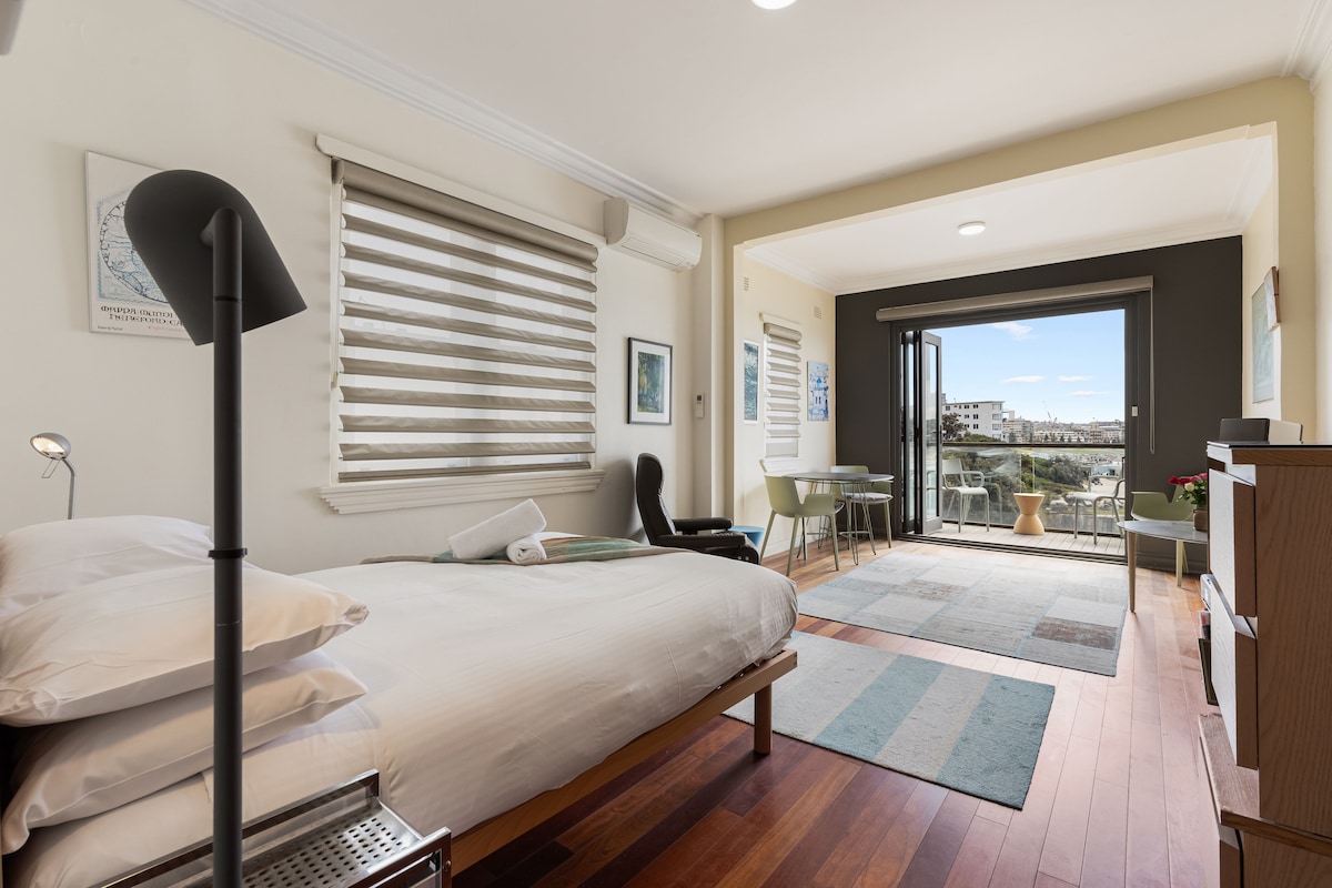 A bright bedroom features a spacious bed with neatly arranged pillows, accompanied by a minimalist lamp and a small side table. A large glass door opens to a private balcony, offering views of the surrounding area. Natural light fills the room from the window with layered blinds.