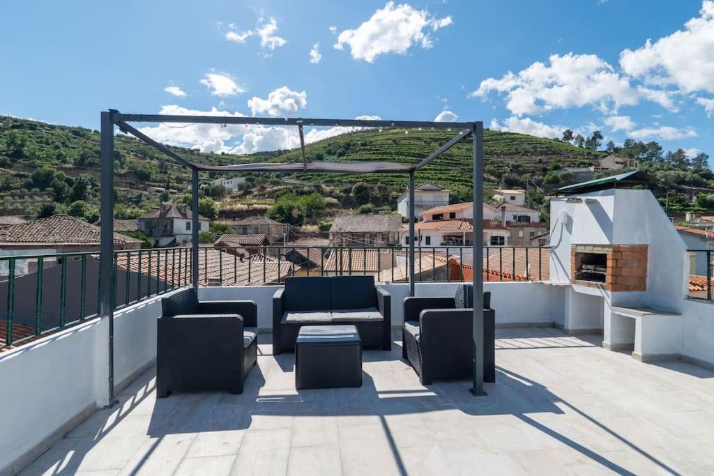 A rooftop terrace offers a view of the surrounding countryside, featuring a seating area with dark wicker furniture. A barbecue grill is positioned nearby, and the landscape is marked by lush greenery and quaint village rooftops under a clear blue sky.