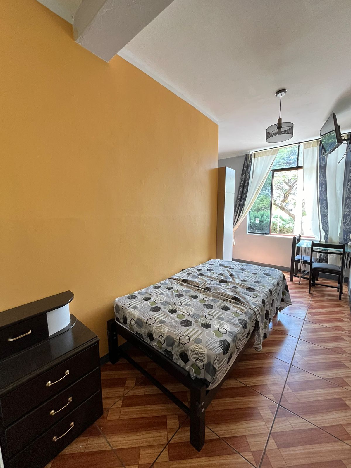 A bright room with an orange accent wall features a single bed covered in a patterned duvet. Natural light enters through a large window, complemented by light curtains. A small desk and chair are positioned nearby, along with a sleek dark dresser.