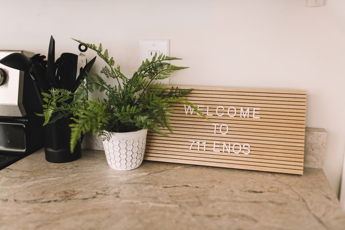 A cozy kitchen corner features a welcome sign made of light wood with white lettering. A small white pot containing a lush fern is placed beside various kitchen utensils in a black container, all set against a textured stone countertop.