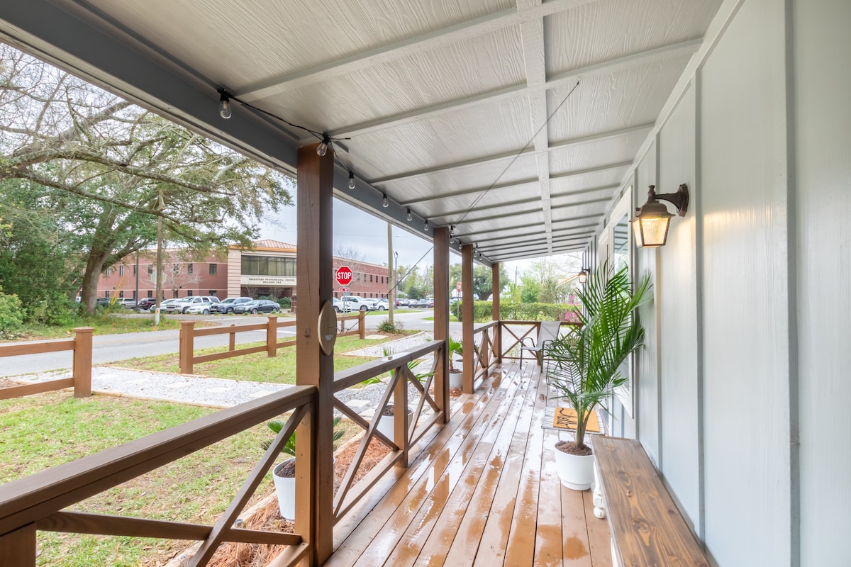 The covered porch features a wide wooden floor, allowing for a comfortable outdoor space. Potted plants are positioned along the railing, and string lights add subtle illumination. A nearby road view reveals parked vehicles and greenery in the distance, enhancing the inviting atmosphere.