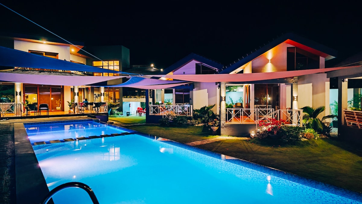 A serene nighttime view of a well-lit swimming pool, reflecting the soft glow of surrounding lights. The pool area is framed by two modern cabins, each displaying large glass windows. Potted plants and shaded seating areas enhance the inviting atmosphere of the outdoor space.