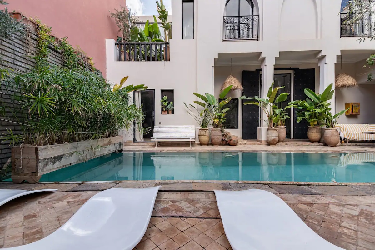 A serene patio is shown, featuring a rectangular swimming pool surrounded by lush greenery. White lounge chairs rest on the tiled deck beside the water. The adjoining walls are adorned with plants in pots, enhancing the tranquil ambiance of the space.