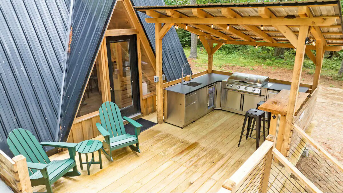 An outdoor kitchen area features stainless steel appliances, including a grill and sink, under a wooden canopy. Two green adirondack chairs are positioned invitingly beside a bar stool on a spacious wooden deck, surrounded by lush greenery.