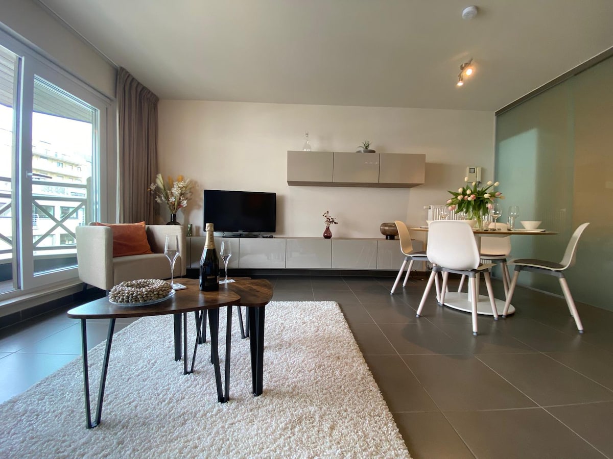 A modern living area features a light-colored area rug beneath a wooden coffee table and a comfortable seating arrangement. A dining table is set for four, accompanied by stylish chairs. Large glass doors allow natural light to fill the space.