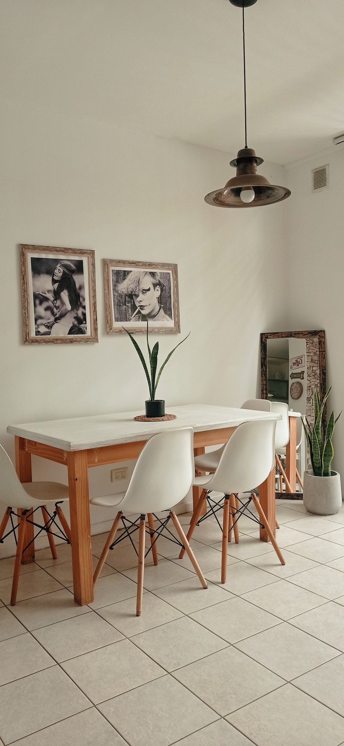 A modern dining area is showcased with a light wood table surrounded by four white chairs. A decorative plant sits at the center of the table. Two framed black-and-white photographs adorn the walls, and a tall plant is positioned next to a large mirror.