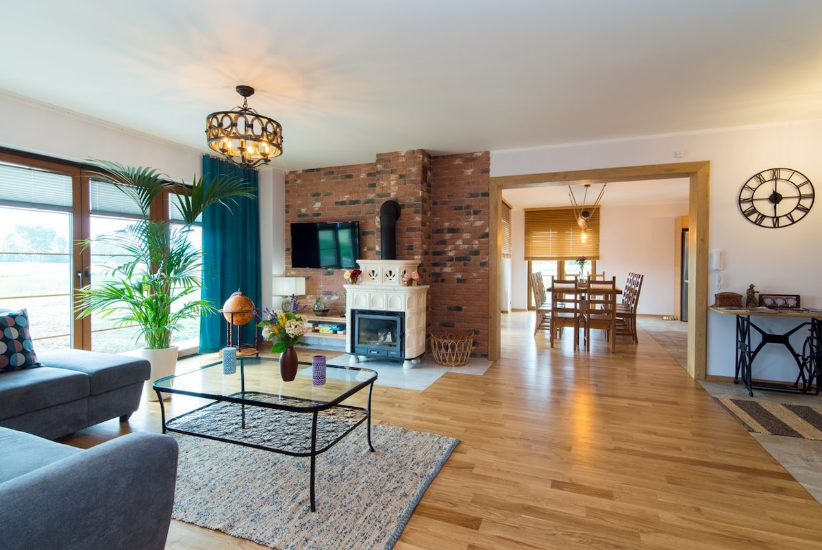 An open living area showcases a comfortable seating arrangement with a sofa and a coffee table set on a textured rug. A bright fireplace feature adds warmth, while large windows invite natural light. A dining space is visible in the background, surrounded by wooden decor.