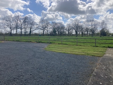 A peaceful outdoor area features a gravel surface leading to a grassy patch. In the background, a grassy field is framed by a line of bare trees under a partly cloudy sky, suggesting a serene natural landscape.