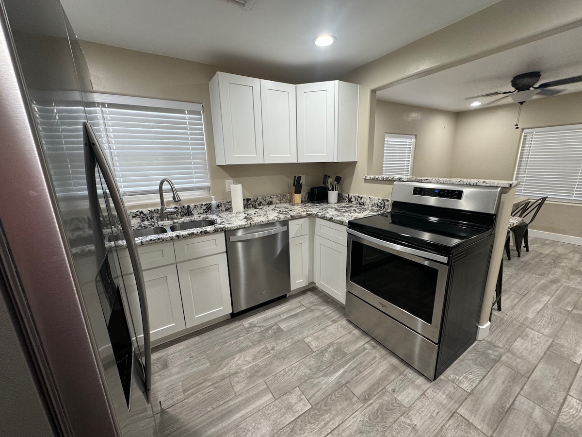 A modern kitchen features white cabinetry and a granite countertop. Stainless steel appliances, including a refrigerator, dishwasher, and stove, are incorporated. Natural light enters through multiple windows, illuminating the space. A dining area with wooden chairs is visible in the background.