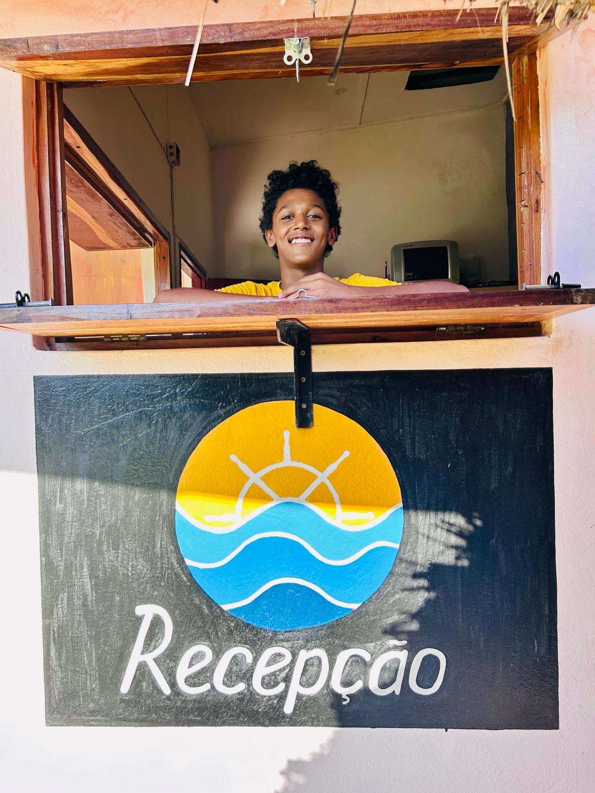 A reception window is visible featuring a colorful sign with a sun and waves motif. The sign displays the word 'Recepção' in white lettering against a black background. Natural light enhances the warm tones of the surrounding area.