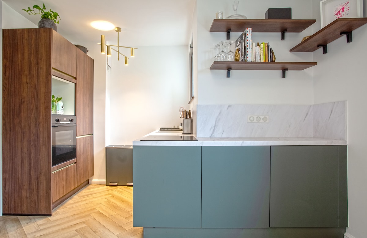 A modern kitchen features sleek cabinetry in muted tones, with a stylish wooden shelving unit displaying books and decorative items. A built-in oven is integrated seamlessly into the cabinetry, complemented by a marble countertop. Natural light enhances the warm tones of the wooden flooring.